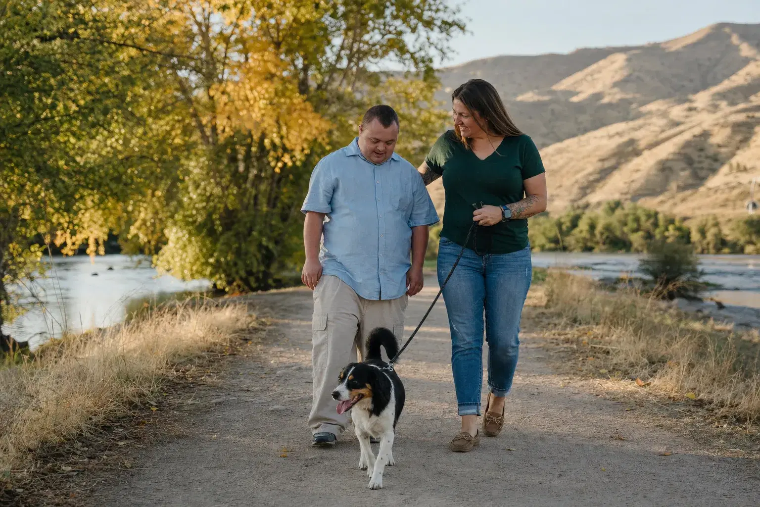 A direct support professional and an adult with a developmental disability walking a dog on a riverside path with the Boise foothills behind them